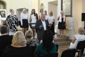 Dyrygent chóru Partytura przemawia do publiczności, za nim członkowie chóru